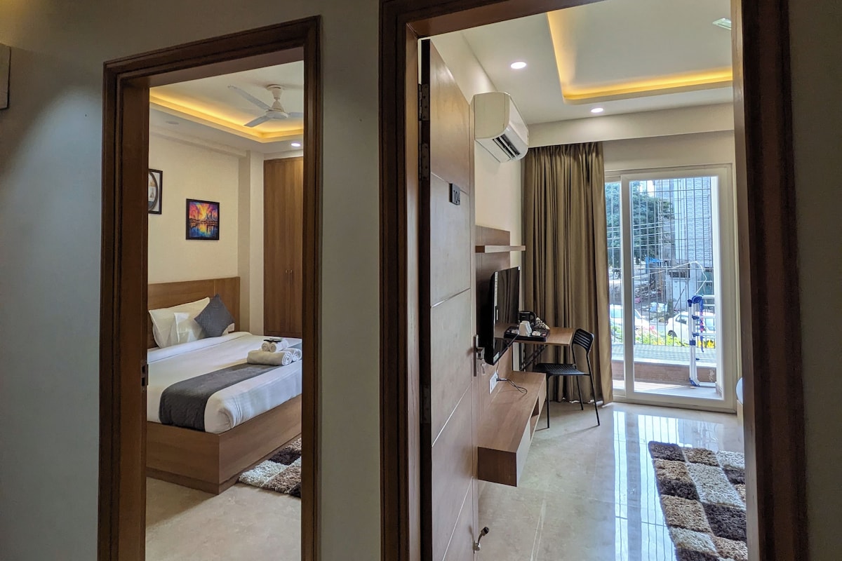 A hotel room with a bed, a desk, a chair, and a view of the city through large windows.