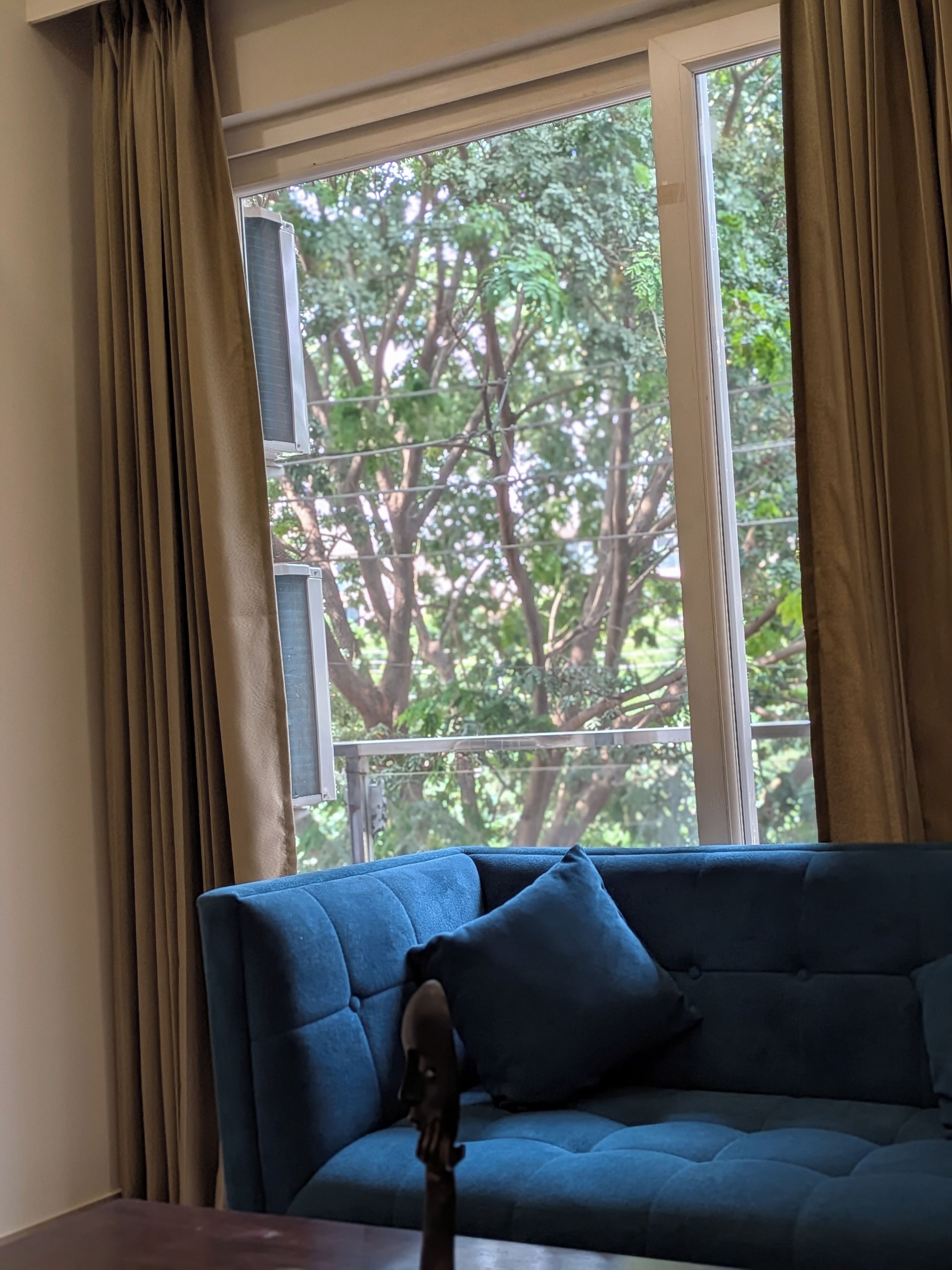 A room with a blue sofa, beige curtains, and a window with a view of trees.