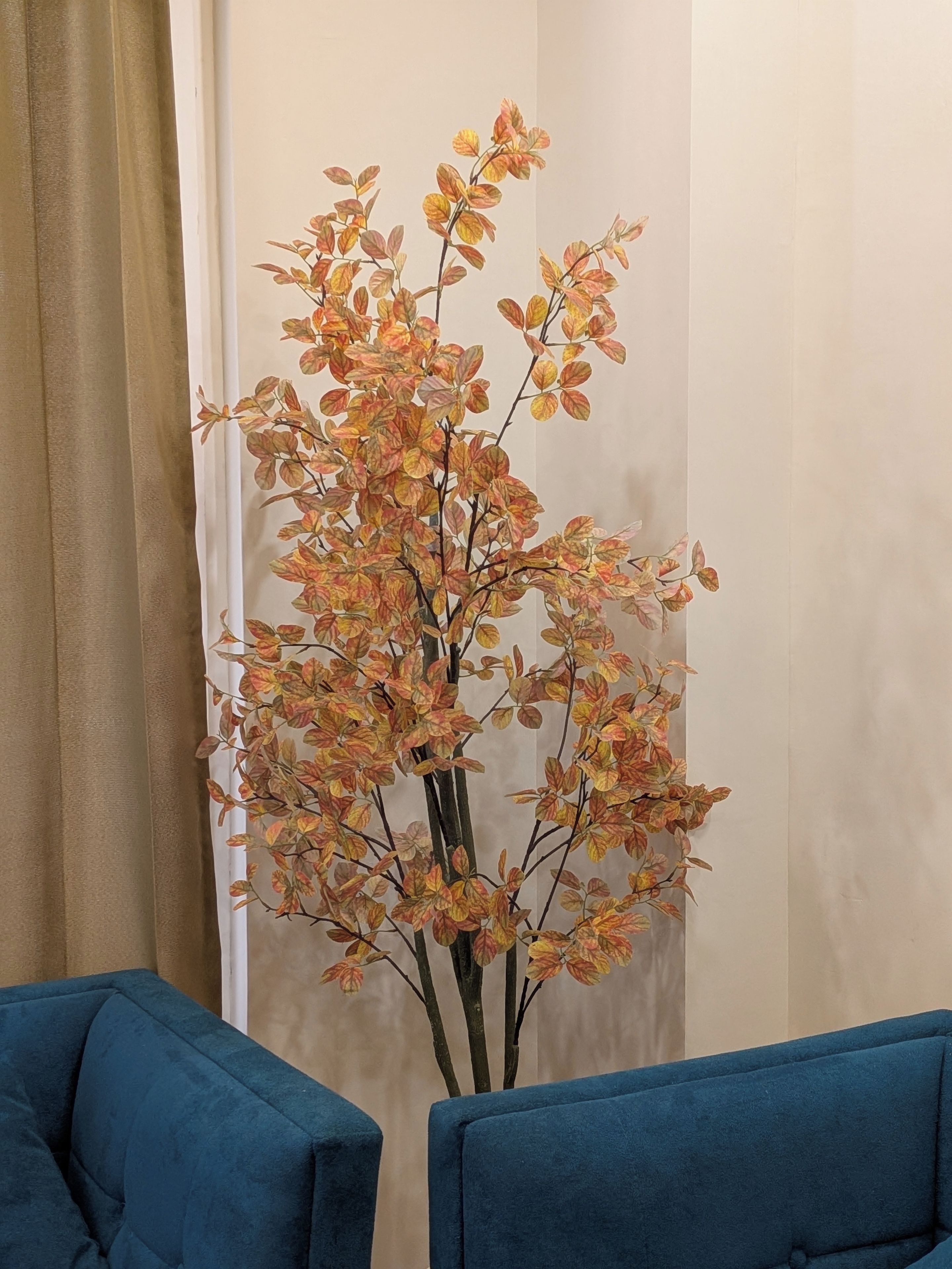 A room with a tall artificial plant in a vase, a blue sofa, and a beige curtain.
