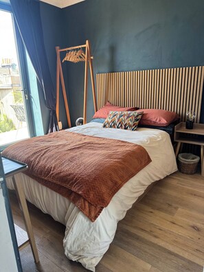 Quarto com cama, cabeceira de madeira, cabideiro, mesa de cabeceira e janela com cortinas.