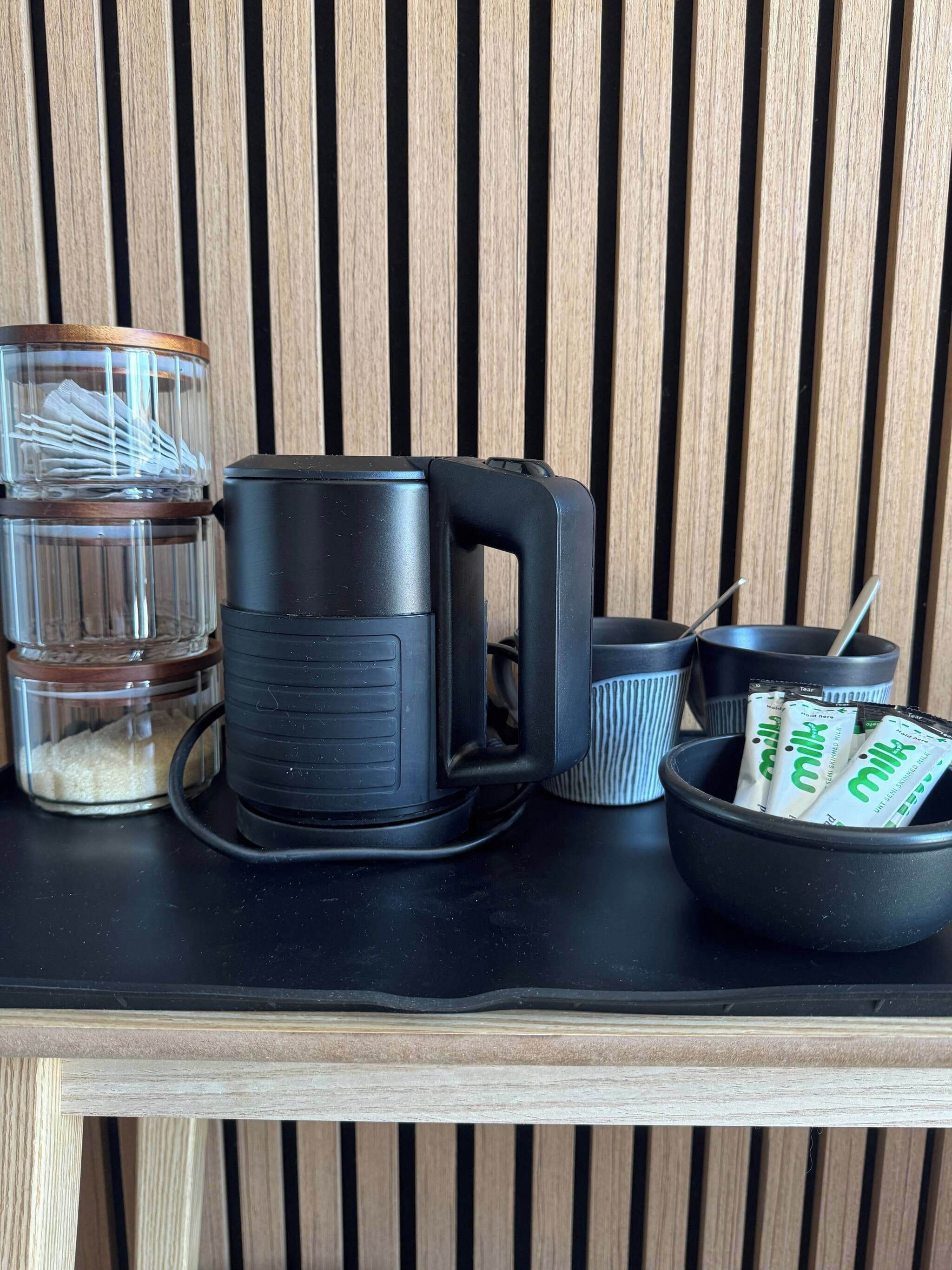 Una encimera de cocina con una cafetera, un recipiente de vidrio con tapa, un tazón con sobrecitos y una taza.