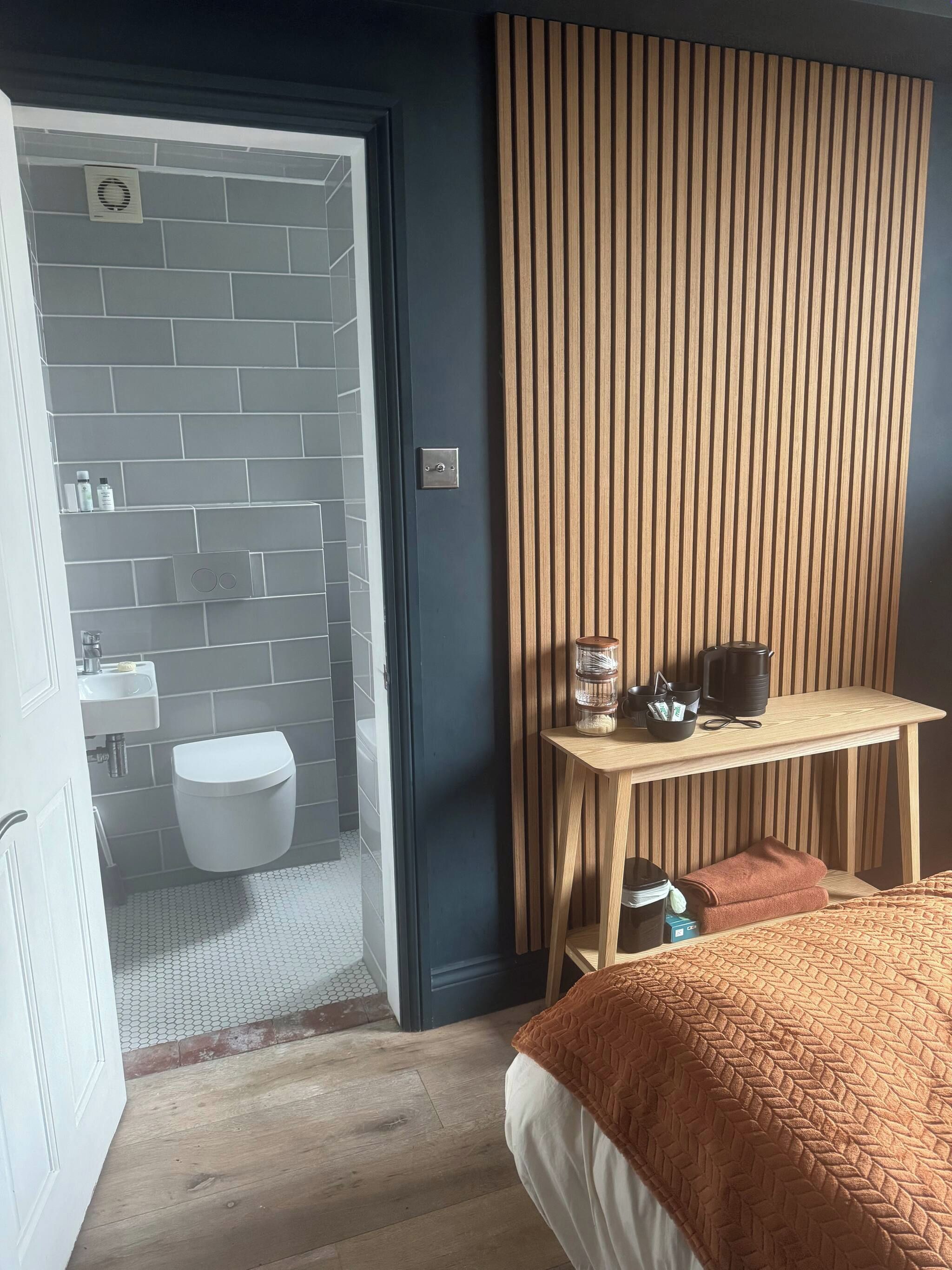 Un baño moderno con inodoro, lavamanos y ducha. Hay un banco de madera con artículos de aseo y una cama con una manta marrón.