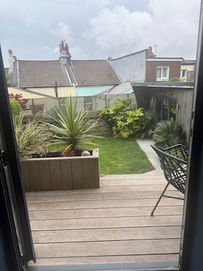 A wooden deck with a planter box containing plants, a pathway leading to a house, and a metal chair.