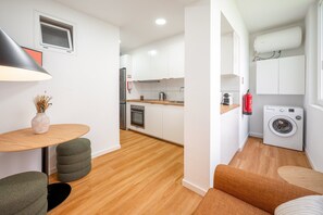 Eine kompakte Küche mit Waschmaschine, ein Essbereich mit Tisch und Stühlen sowie eine kleine Sitzecke mit Sofa und Hocker.