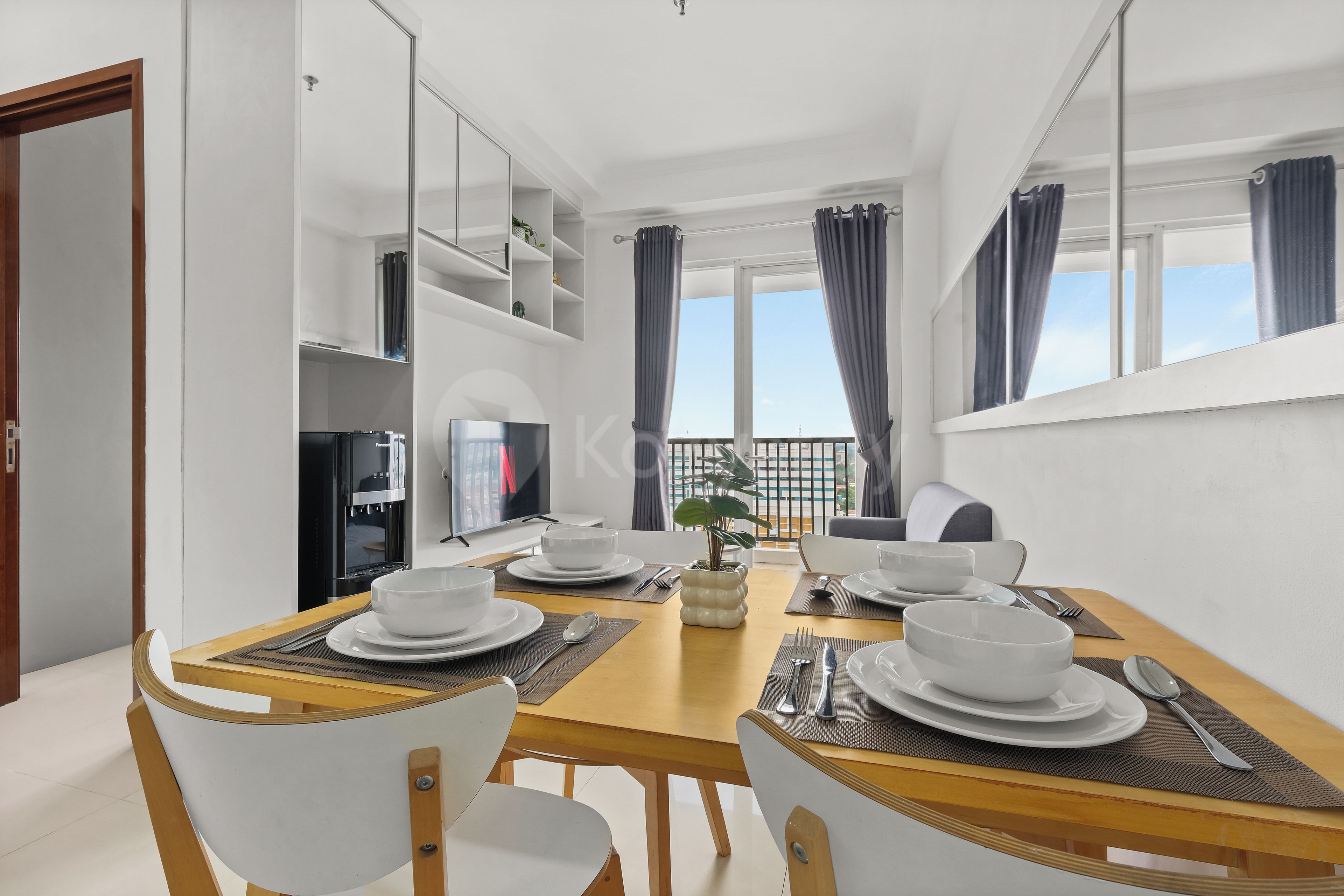 Un espace de repas moderne avec une table en bois dressée pour quatre personnes, des chaises blanches et une vue sur la ville grâce à de grandes fenêtres.
