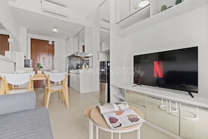 Ein modernes Wohnzimmer mit Flachbildfernseher, einem Essbereich mit Tisch und Stühlen sowie einer Küche mit weißen Schränken und Geräten.