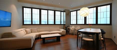 A modern living room with a sectional sofa, a dining table with chairs, and large windows with frosted glass panels.