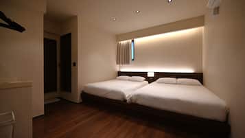A hotel room with two beds, a small window with curtains, a wooden floor, and recessed ceiling lighting.