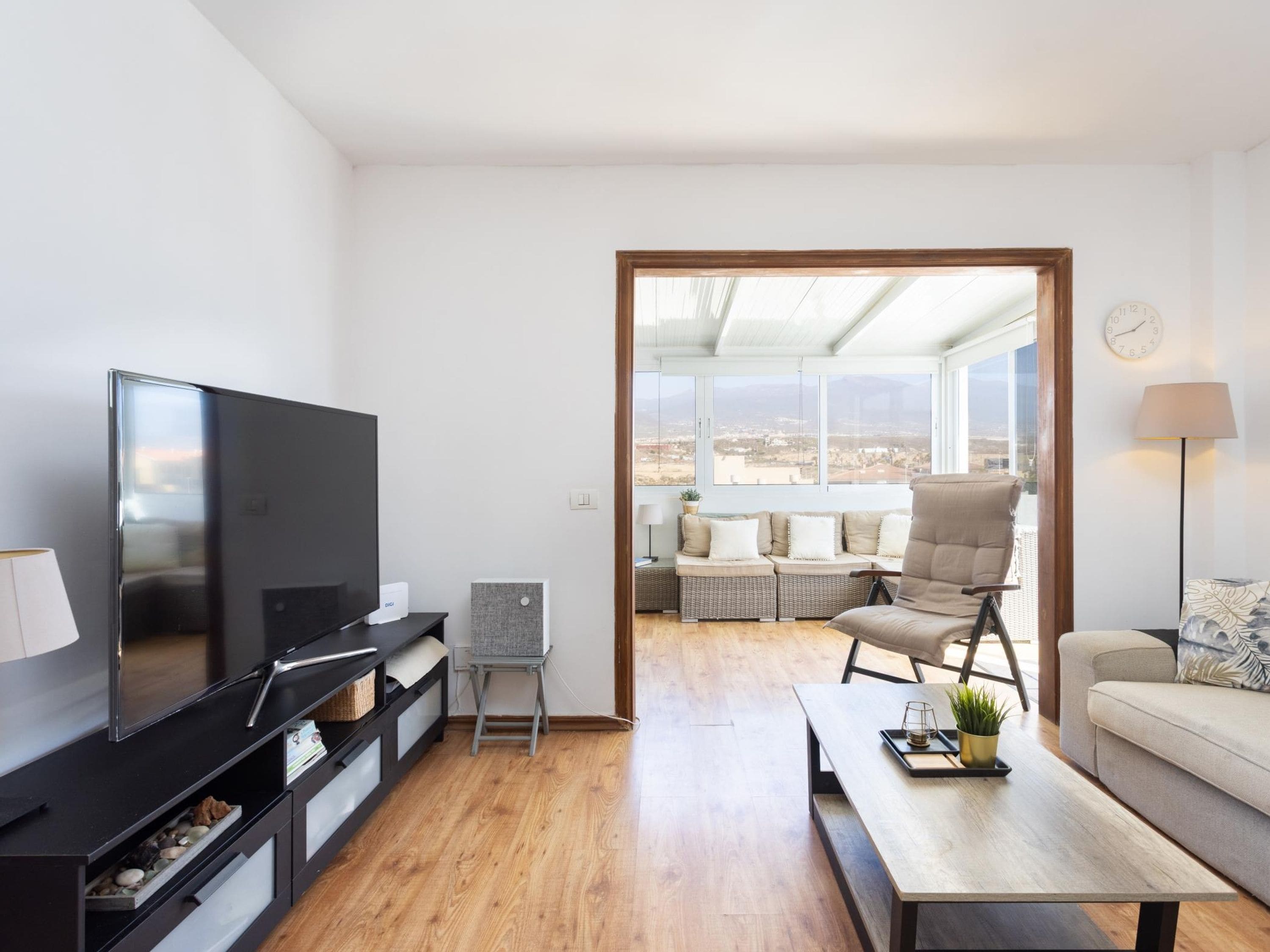 A modern living room with a flat-screen TV, a sofa, a coffee table, and a view through a large window.