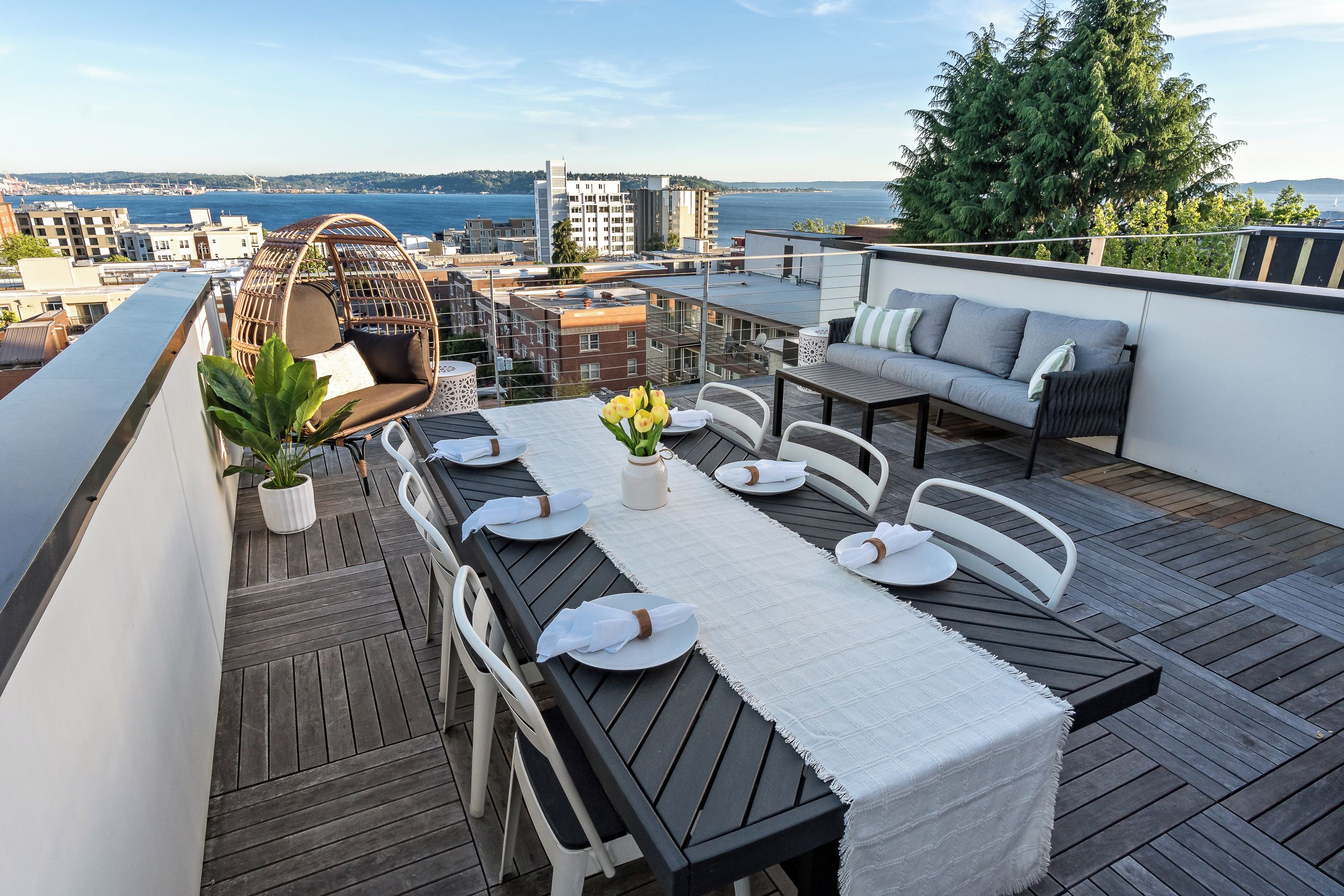 A rooftop terrace with a dining table set for four, surrounded by white chairs, a wicker chair, and a grey sofa.