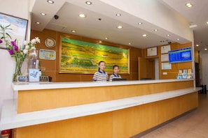 A reception desk with two staff members, a clock, a large landscape painting, and various informational posters. - 7 Days Inn (Miyun)