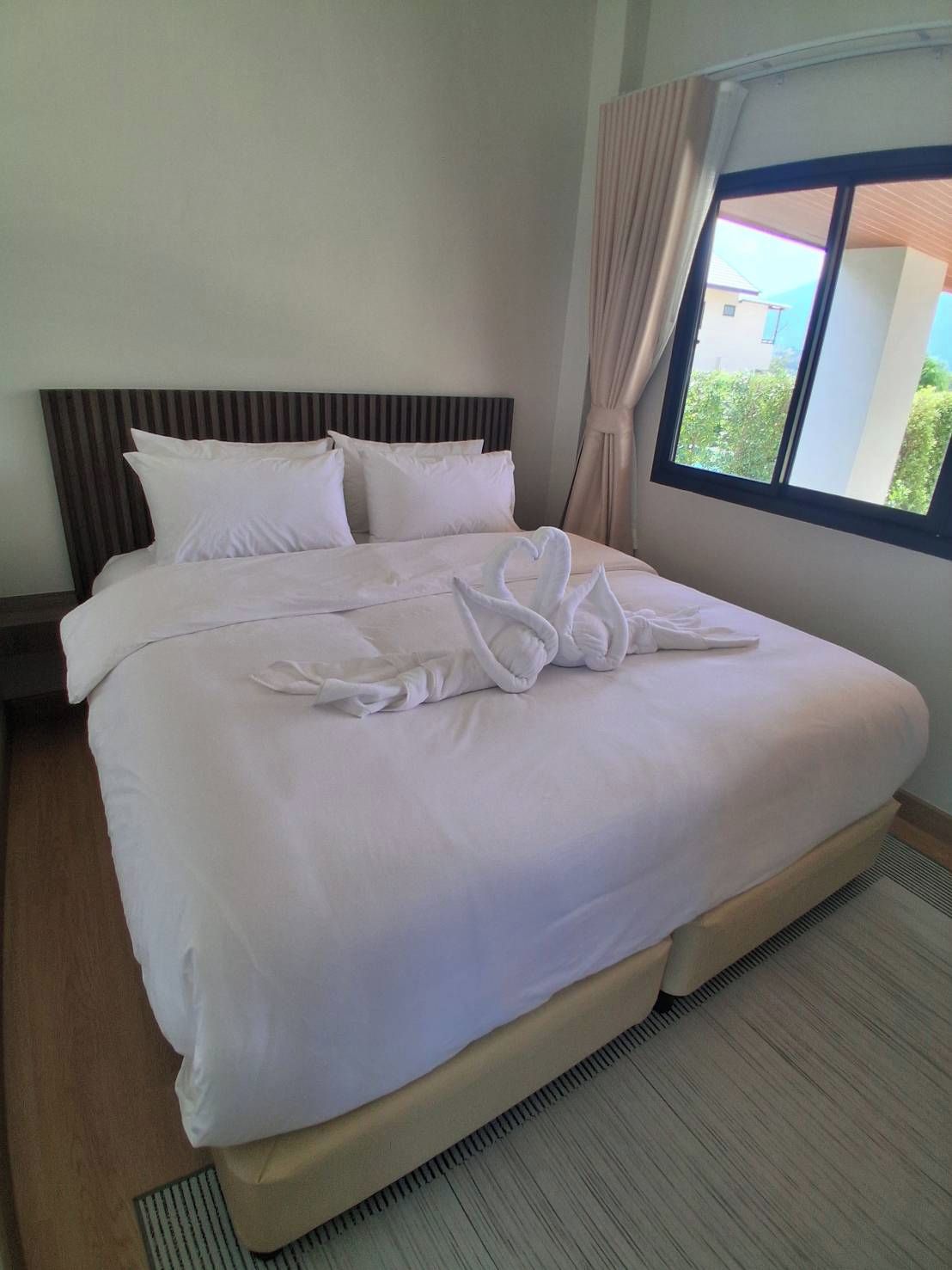Habitación de hotel con una cama grande, cabecera, ventana con cortinas y vistas al exterior con vegetación.