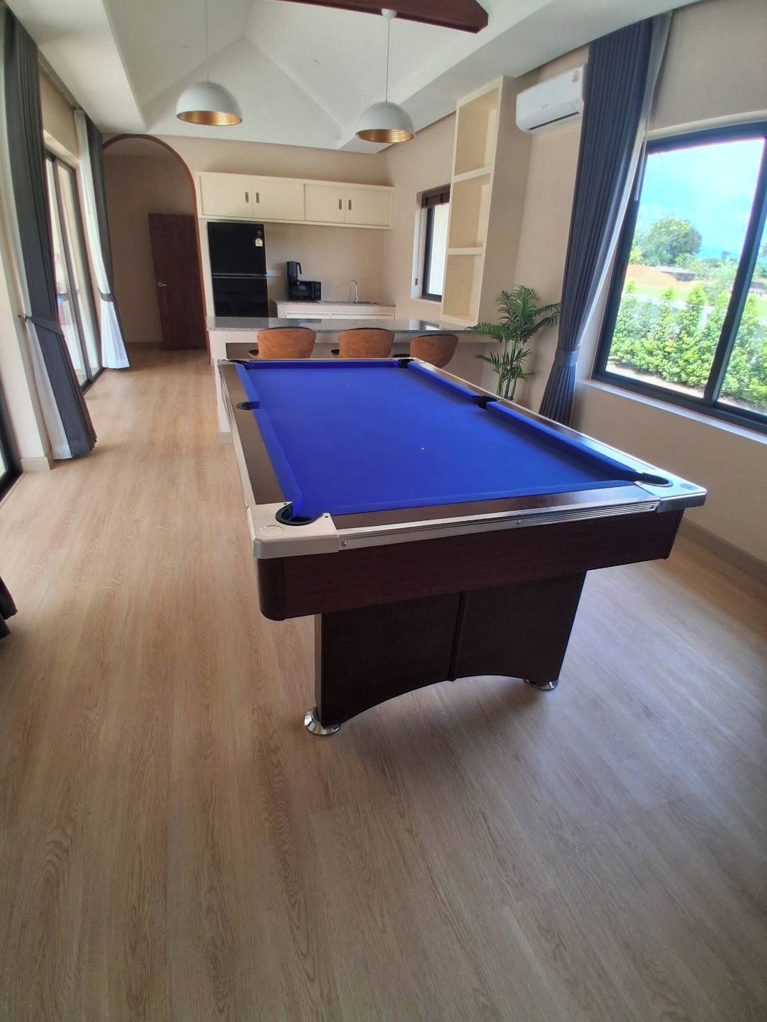 A pool table in a room with a kitchen area, large windows, and a view of greenery outside.