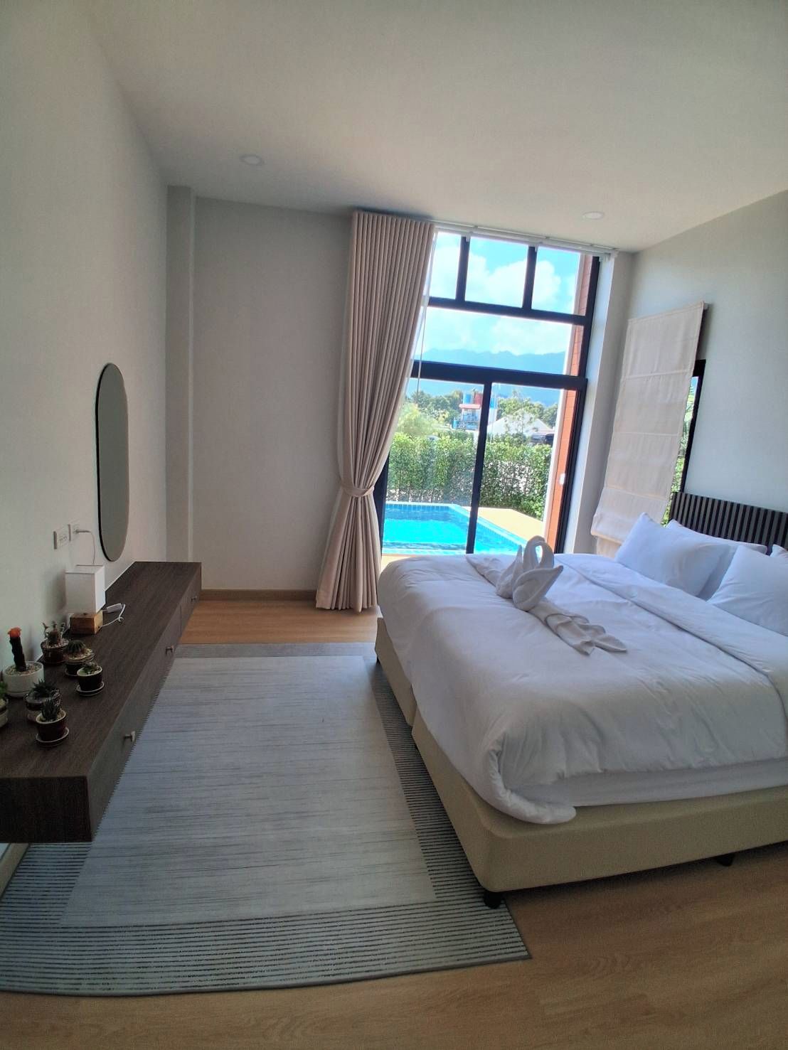 A hotel room with a large bed, a bedside table with a mirror, a small potted plant arrangement, a view of a pool and mountains through the window.