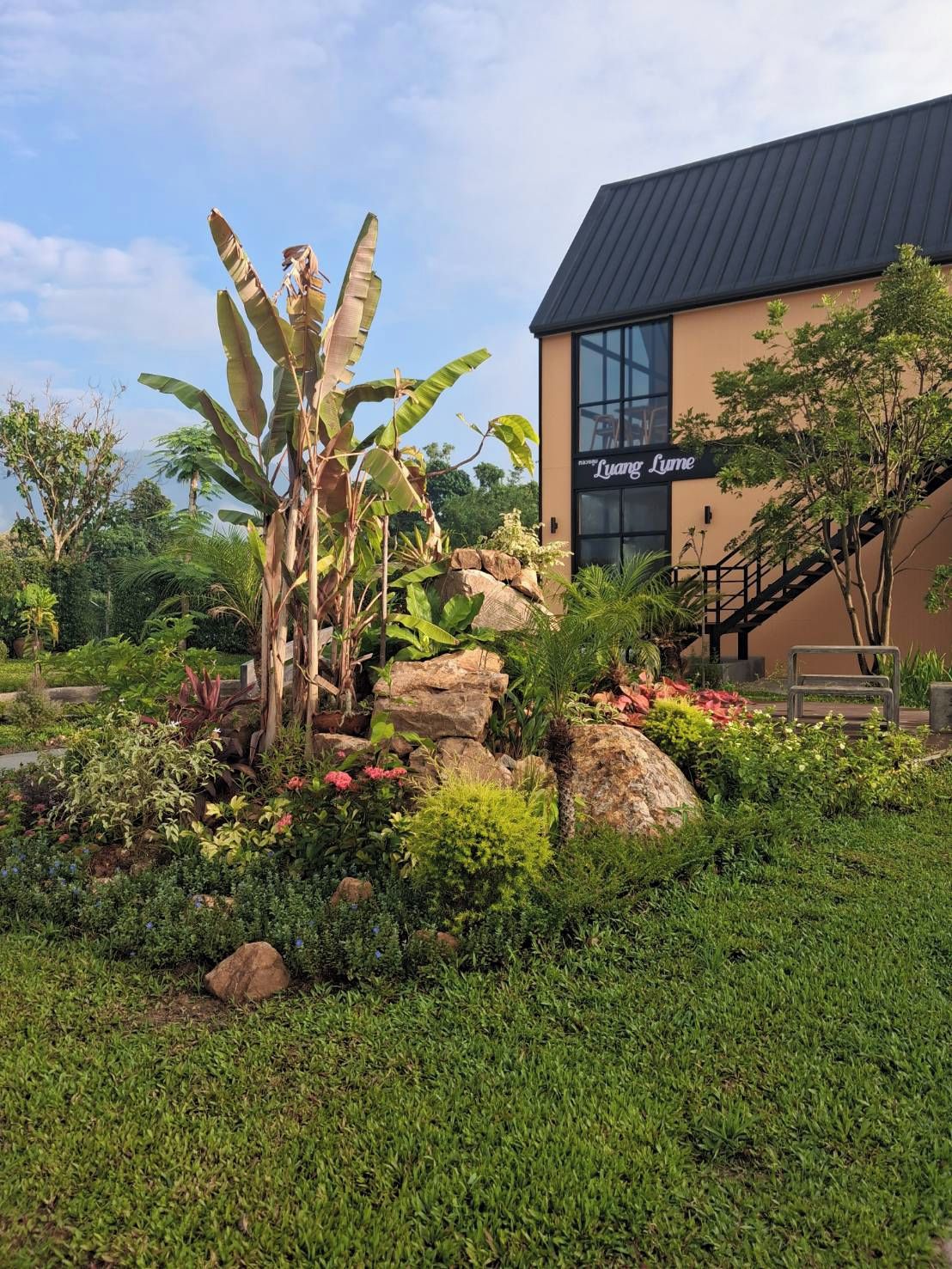 A building with a sign reading 'Luang Lum' surrounded by a landscaped garden with rocks, plants, and a banana tree.