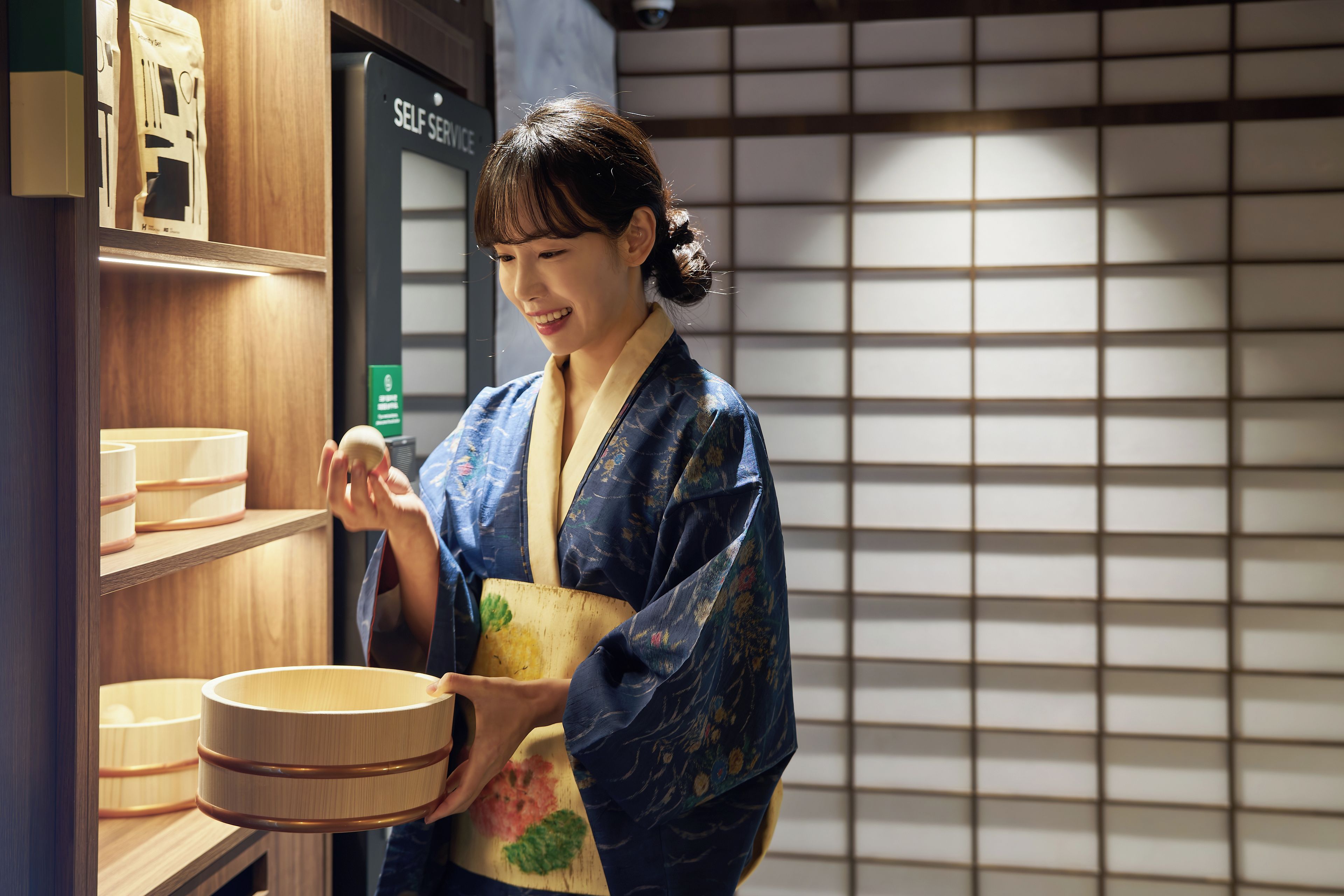A person in traditional attire holding a wooden object, with shelves and a sign labeled 'SELF SERVICE' in the background.