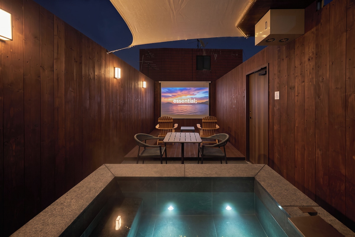 Un área de piscina con paredes de madera, una mesa con sillas y una pantalla de proyección que muestra 'essential' sobre un fondo pintoresco.