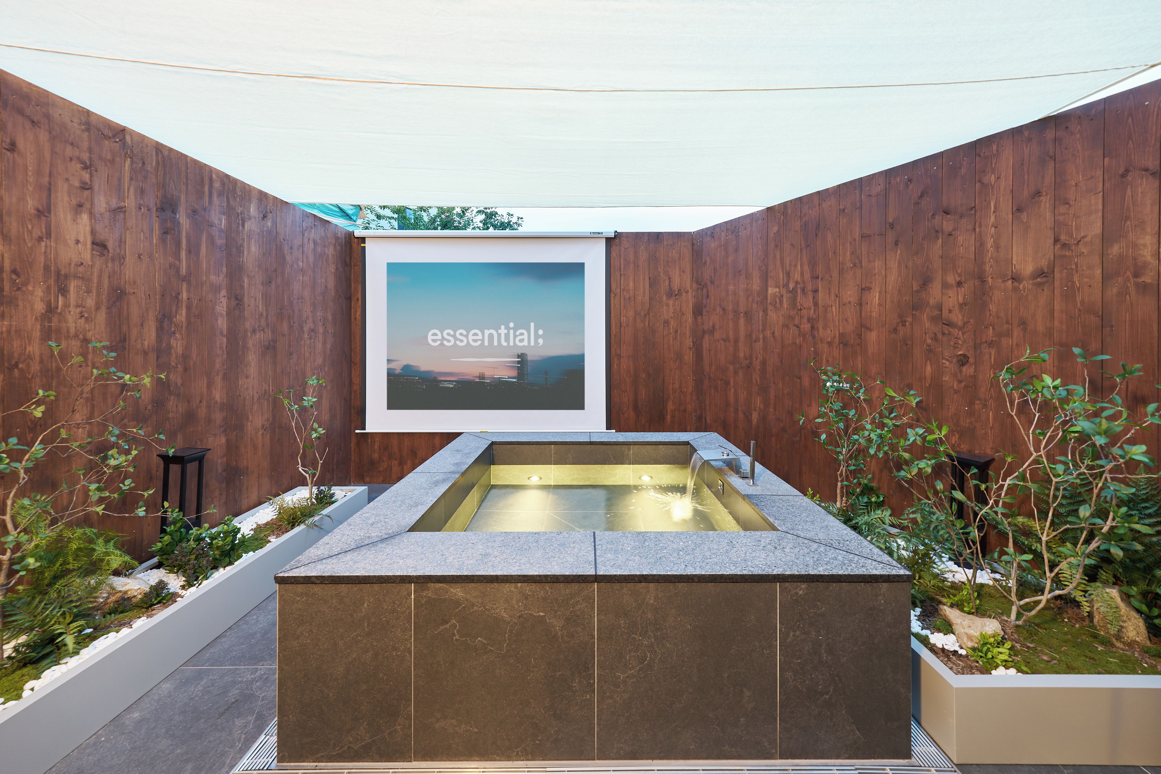 A modern outdoor spa area with a rectangular hot tub, wooden walls, and a framed picture with the word 'essential' on it.