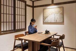 A person seated at a wooden table with a teapot and cups, in a room with traditional Japanese sliding doors and a framed artwork on the wall.