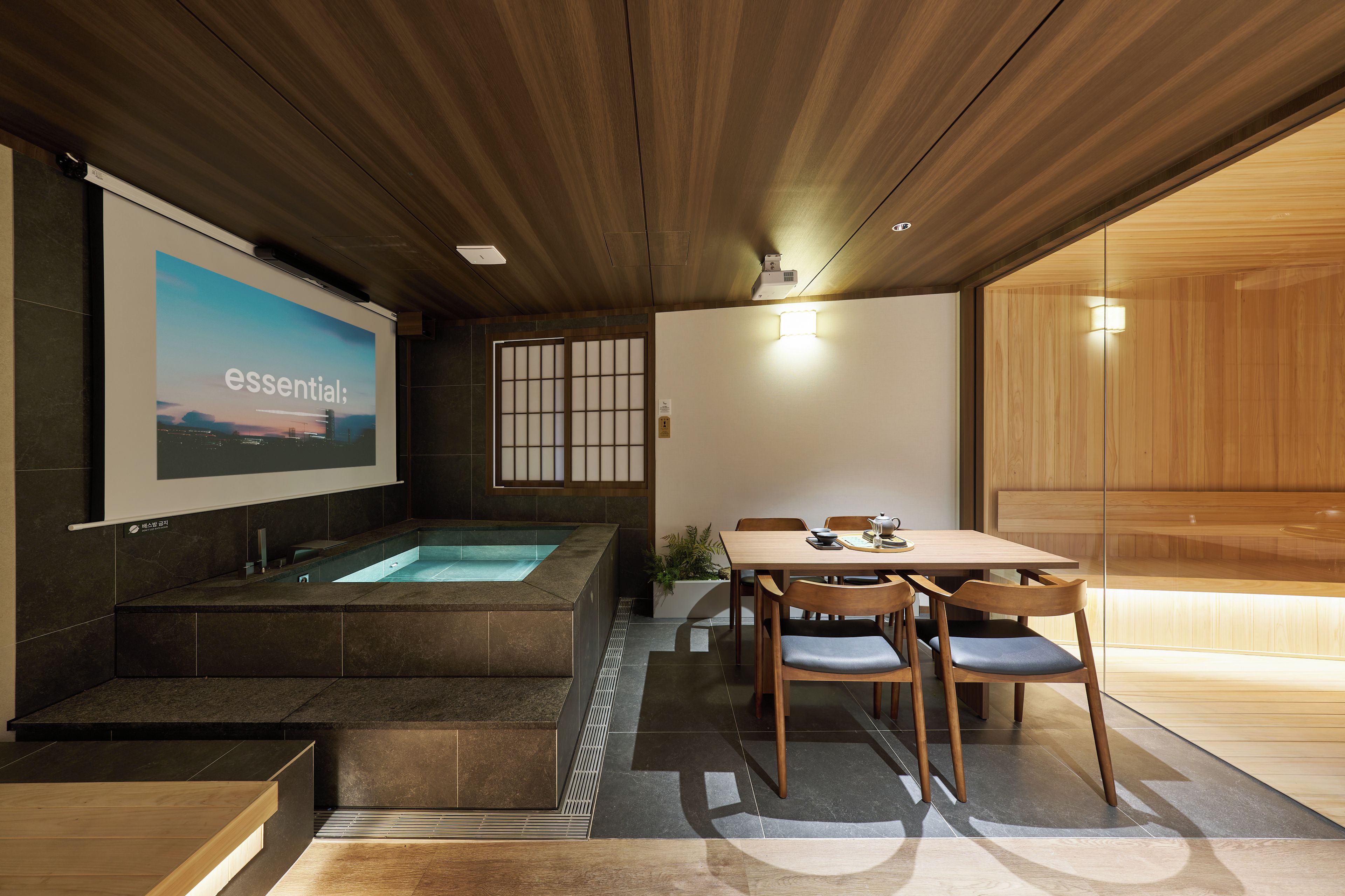 Un interior moderno con techo de madera, una pantalla grande mostrando 'essential', un jacuzzi y un comedor con sillas y mesa.