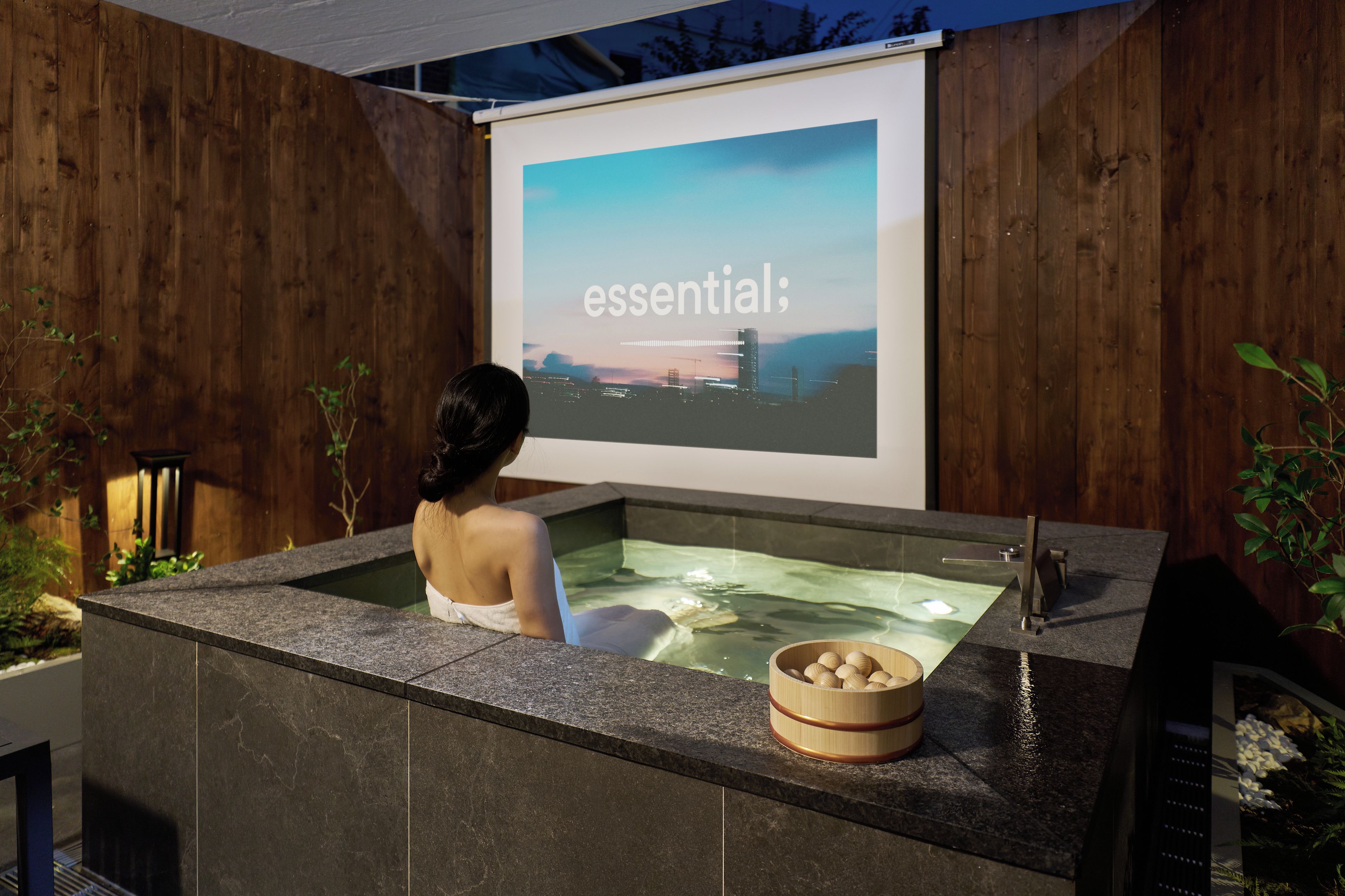 A person relaxing in a hot tub with a projection screen displaying the word 'essential' in the background.