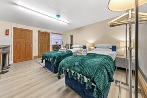 A hotel room with two beds, a bedside table, a lamp, a window with curtains, a fire extinguisher, and a door. - Cozy East Ham Studio | 2 Double Beds | Sleeps 4 (Greater London)