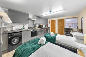 Ein modernes Hotelzimmer mit einer Küchenzeile, einer Waschmaschine, zwei Betten mit grünen Decken, einem Fernseher und einem Fenster mit Blick.