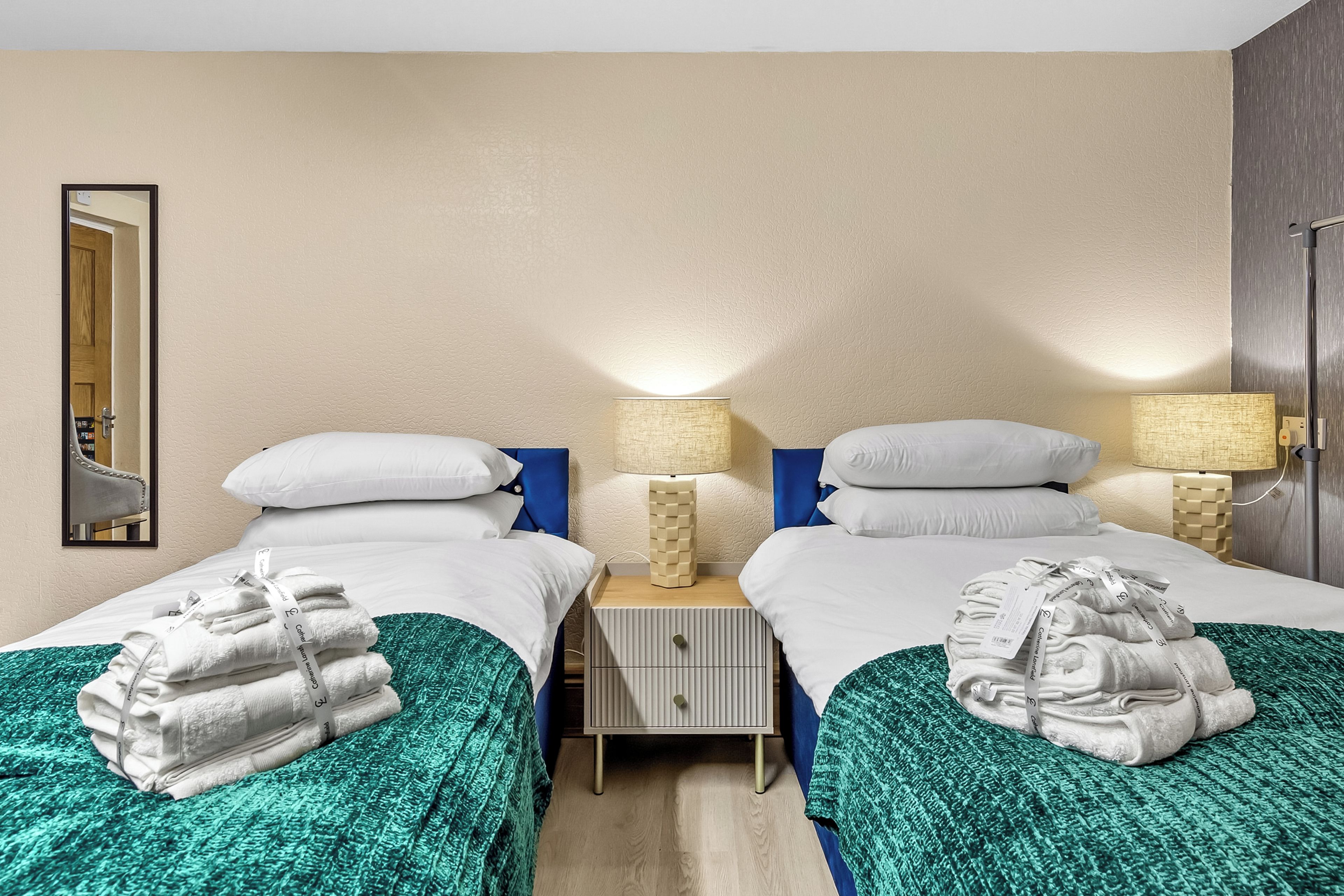 A hotel room with two beds, each with white linens and a green blanket. There are two bedside tables with lamps, a mirror on the wall, and a door leading to another room.