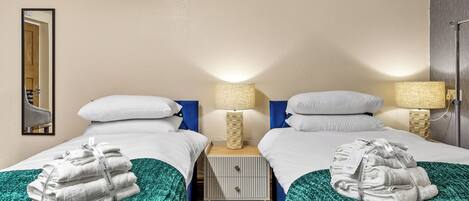 A hotel room with two beds, each with white linens and a green blanket. There are two bedside tables with lamps, a mirror on the wall, and a door leading to another room.