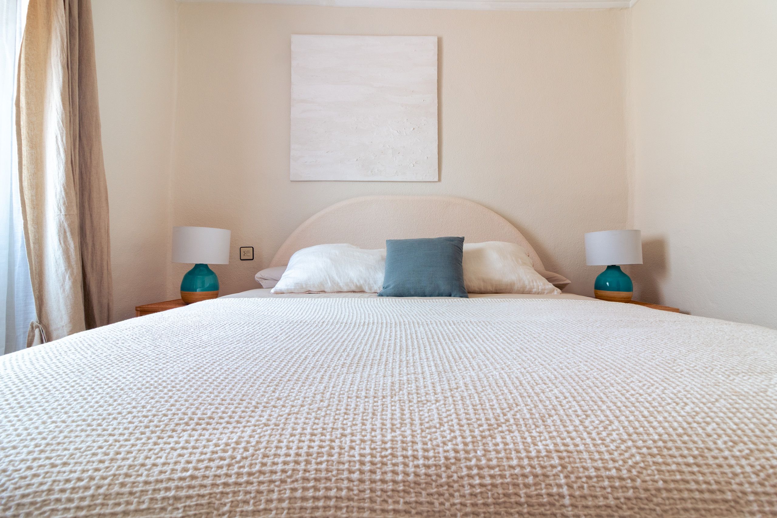 Un lit bien fait, recouvert d’une couette blanche à texture intéressante, avec deux oreillers et un seul oreiller bleu. On y trouve deux tables de chevet sur lesquelles sont posées des lampes de couleur turquoise.