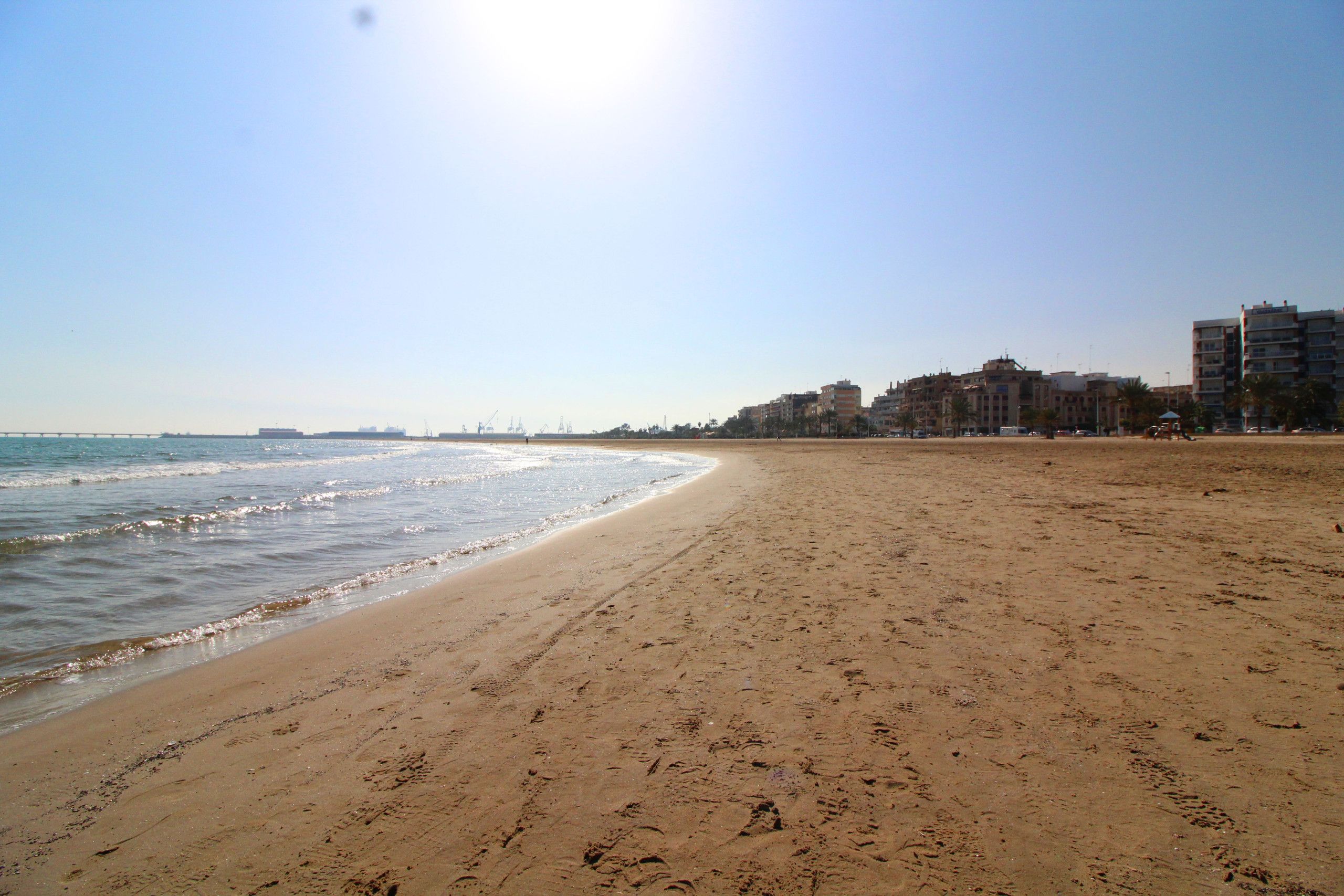 Une vaste plage de sable avec des empreintes, une mer calme et des bâtiments en arrière-plan.