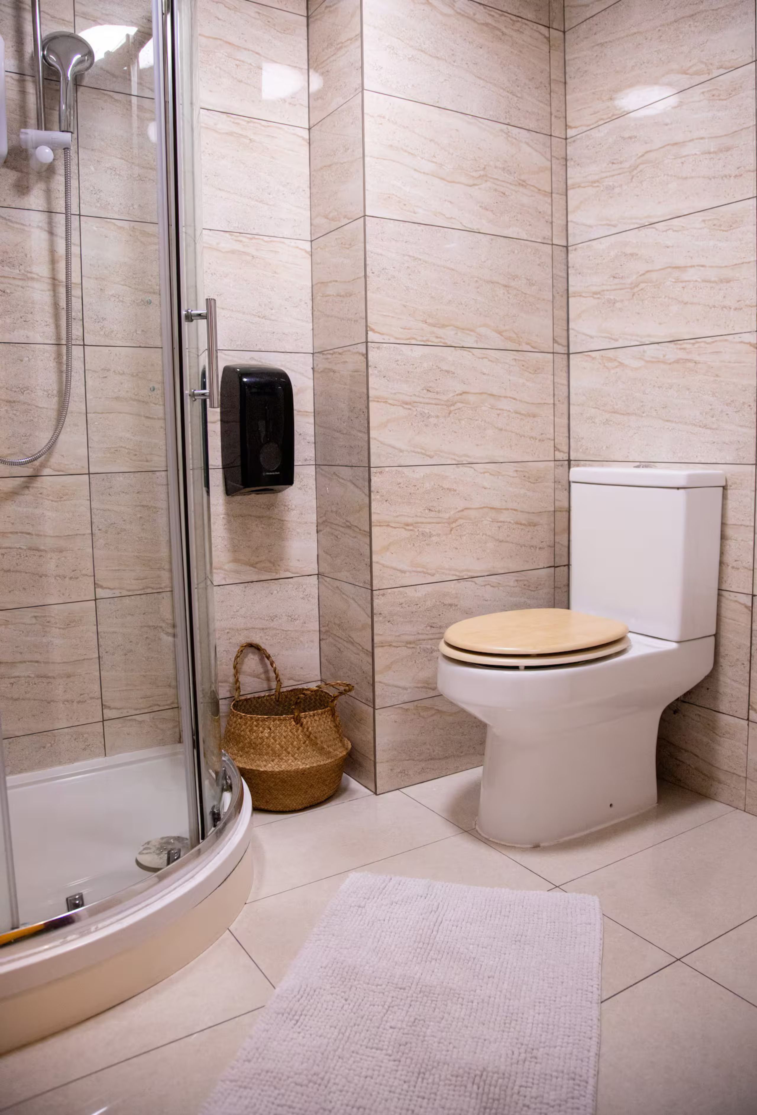 A bathroom with a glass shower enclosure, a toilet with a wooden seat, a hand dryer, and a woven basket.