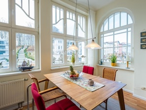 A dining area with a wooden table, red chairs, and large windows offering a view of buildings and a street. - Holiday Apartment With 2 Bedrooms (Sellin)