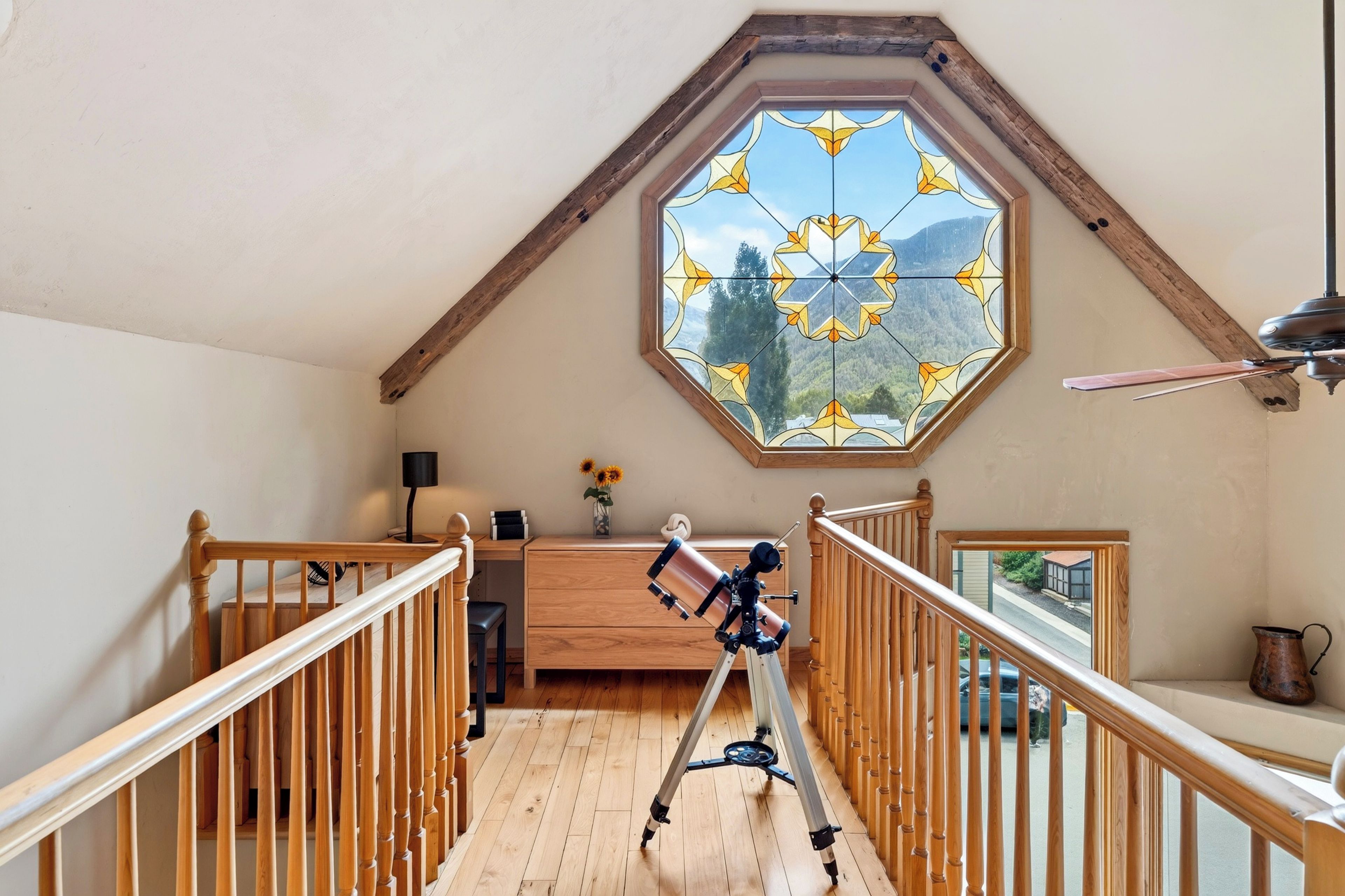Une pièce avec un télescope posé sur un plancher en bois, un bureau en bois, une lampe et une fenêtre donnant sur une vue sur les montagnes.