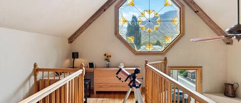 Une pièce avec un télescope posé sur un plancher en bois, un bureau en bois, une lampe et une fenêtre donnant sur une vue sur les montagnes.