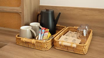 A wicker tray with a kettle, mugs, coffee packets, and sugar packets on a wooden floor.
