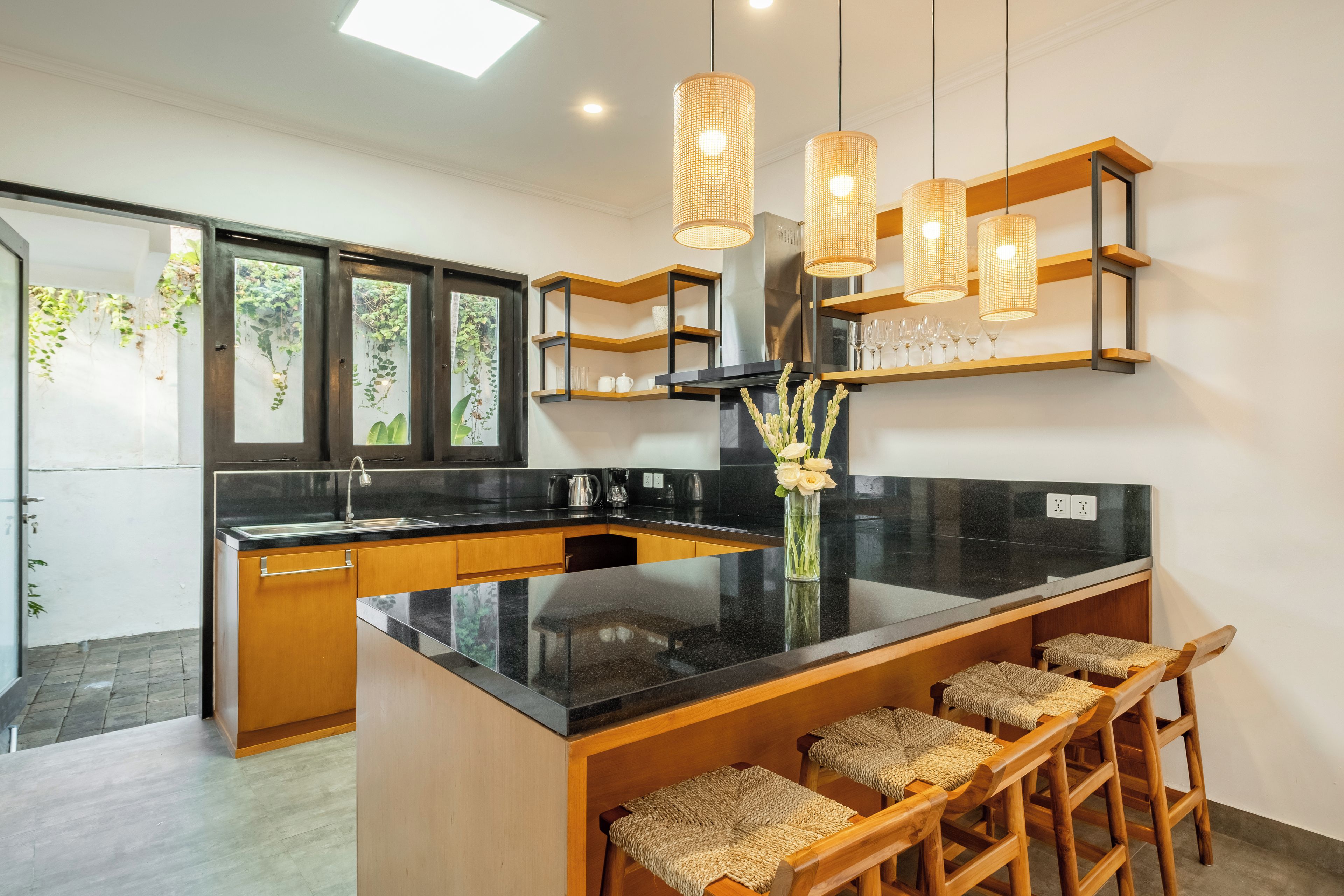 Une cuisine moderne dotée d’un îlot central, de tabourets de bar en bois, de luminaires suspendus et offrant une vue sur la verdure environnante.