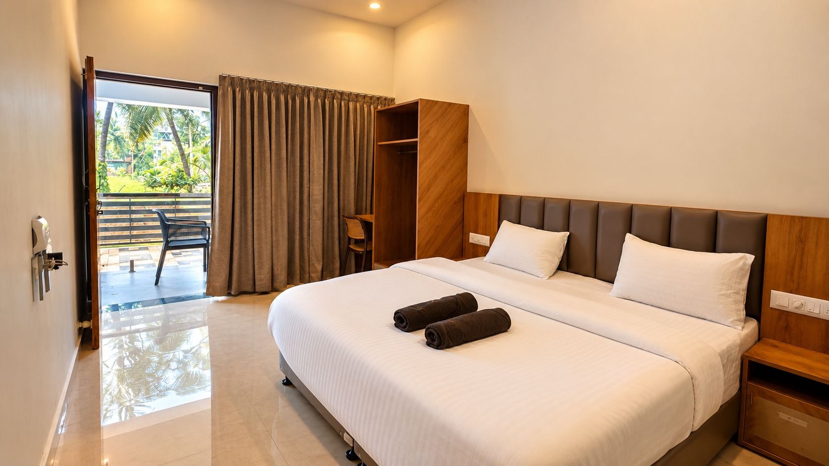 Habitación de hotel con una cama, cabecera de madera, mesita de noche y vistas a un balcón con una silla y cortinas.