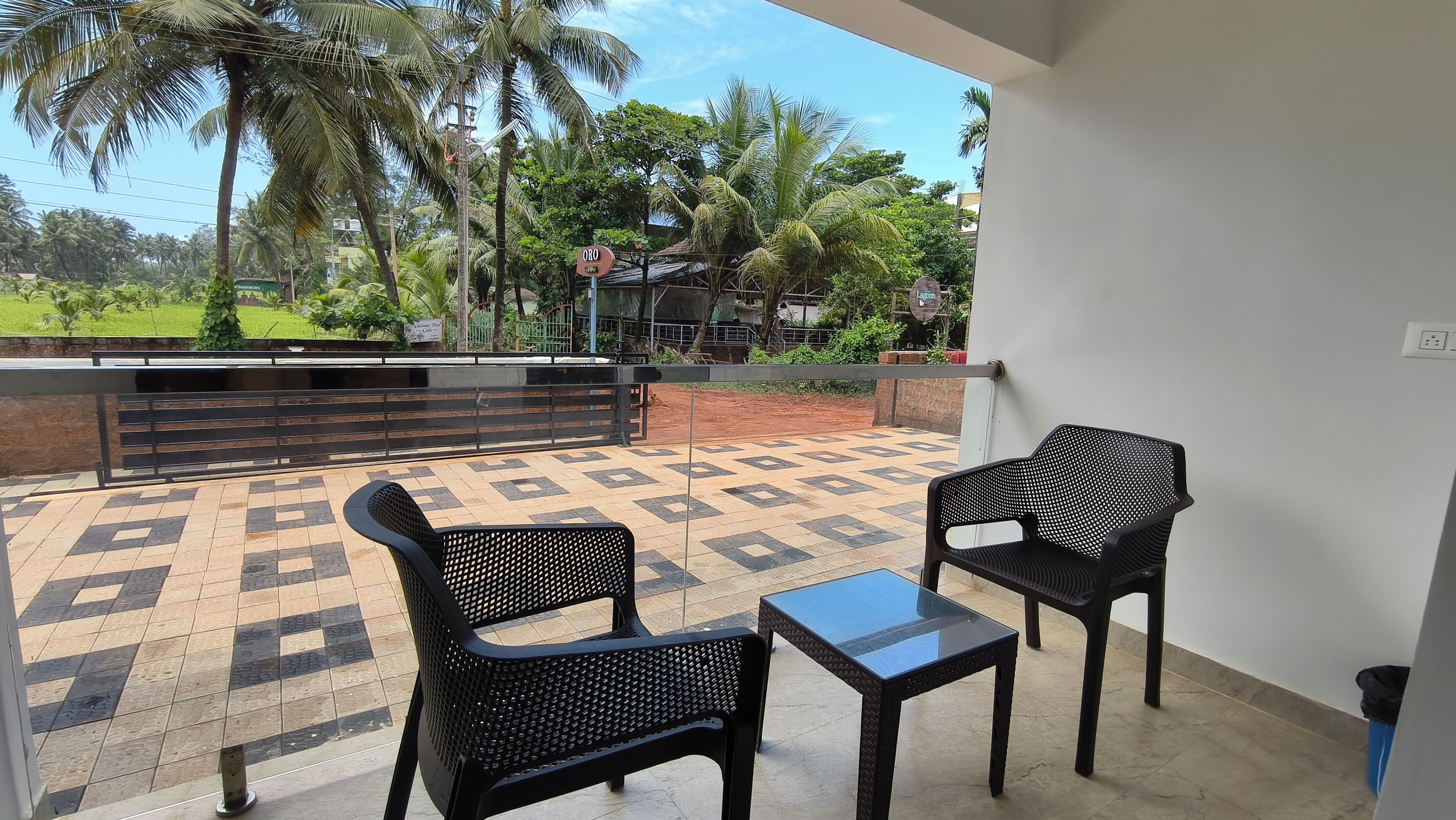 Un balcón con dos sillas negras y una pequeña mesa, con vistas a un jardín con palmeras y un edificio al fondo.