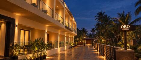 Un hotel con diseño moderno, que cuenta con un amplio espacio exterior con pasarela empavedada, plantas en macetas y palmeras.