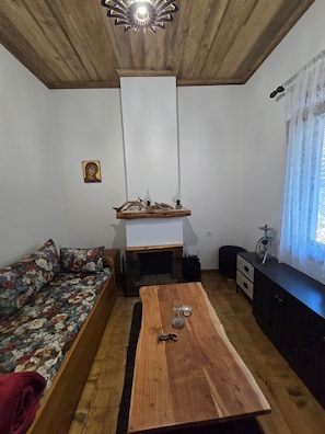 A room with a wooden ceiling, a sofa with floral upholstery, a wooden coffee table, a small fireplace, and a window with sheer curtains.