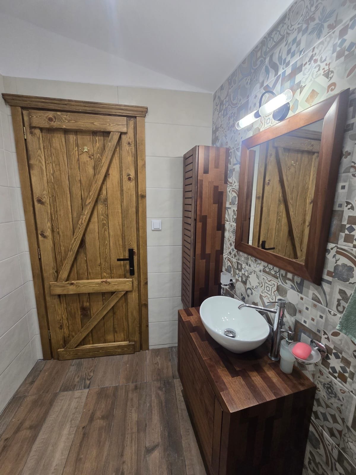A bathroom with a wooden door, a sink with a mirror, and patterned tiles on the walls.