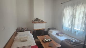 A small, cozy room with a wooden ceiling, a bed with a red blanket, a table with a chessboard, and a window with sheer curtains.