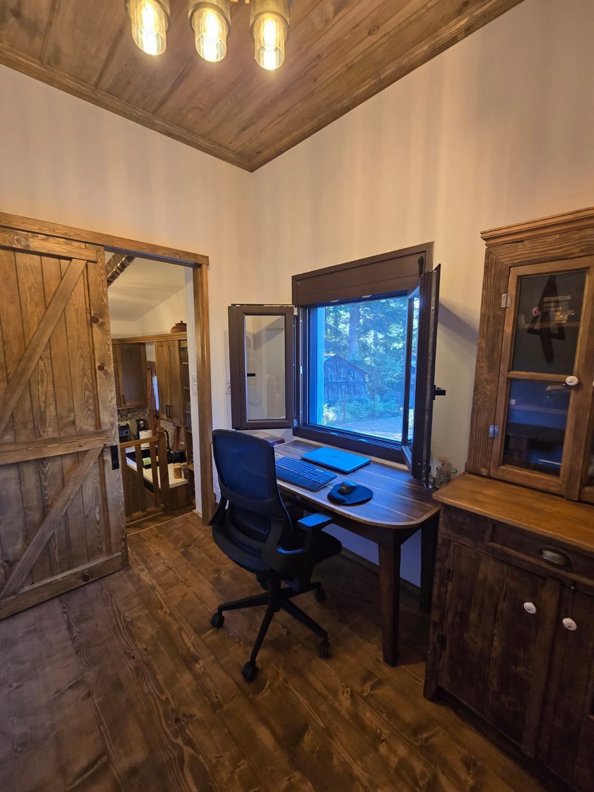 A room with a wooden desk, a computer, and a chair. There is a window with a view of trees and a building outside.