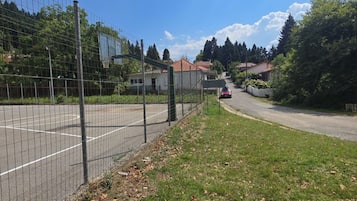 A fenced sports court with a road leading to houses, surrounded by trees and a clear sky.