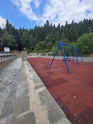 A playground with a swing set and a red rubberized surface, surrounded by a stone wall and a forested hillside. - Oniropagida House in Beautiful Nature (Central Tzoumerka)