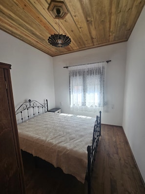 A bedroom with a wooden ceiling, a bed with a metal frame, a window with a sheer curtain, and a small bedside table. - Oniropagida House in Beautiful Nature (Central Tzoumerka)