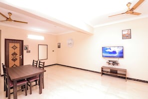 A dining area with a table and chairs, a ceiling fan, a television on a stand, and a wall with a clock and framed pictures. - LUXE LIVING Korattur Near IT Park  (Chennai)