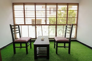 A room with a table and two chairs, a window with a view of buildings, and artificial green carpet.