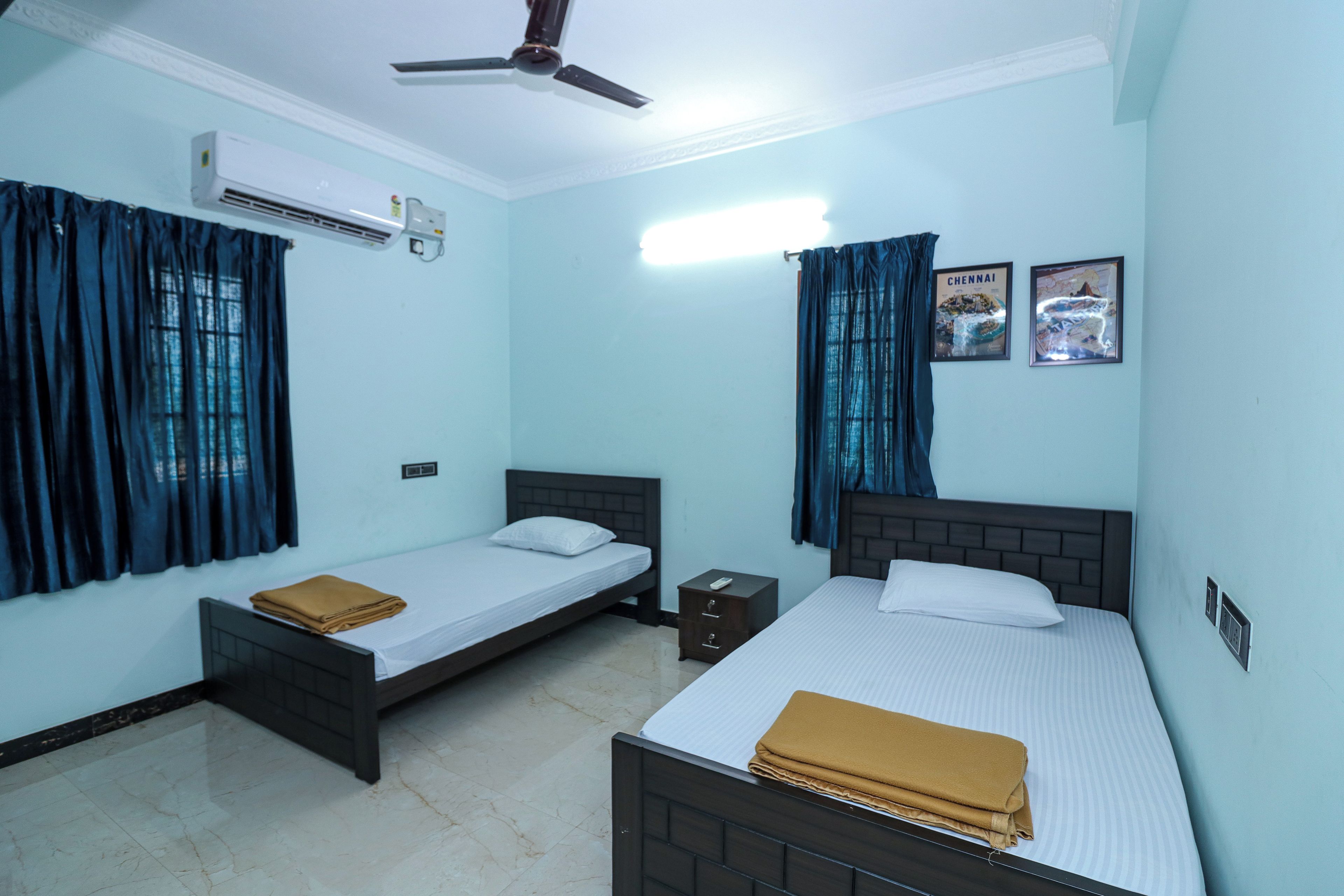 A hotel room with two single beds, a ceiling fan, an air conditioning unit, and two framed pictures on the wall.
